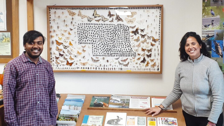 A brown man with dark hair and a beard stands on the left in a purple shirt, across from a brown woman with long brown hair in a grey shirt. They both stand in front of a large entomology display with insects surrounding an illustration in the shape of Nebraska.