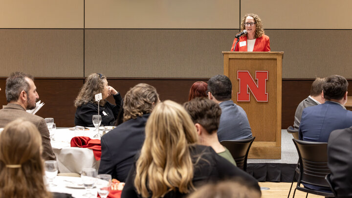 Kate Brooks, head of the University of Nebraska–Lincoln Department of Agricultural Economics, speaks at a podium with a red “N” logo while presenting awards during the department’s Student, Alumni and Service Recognition Banquet as attendees sit at round tables in the audience.